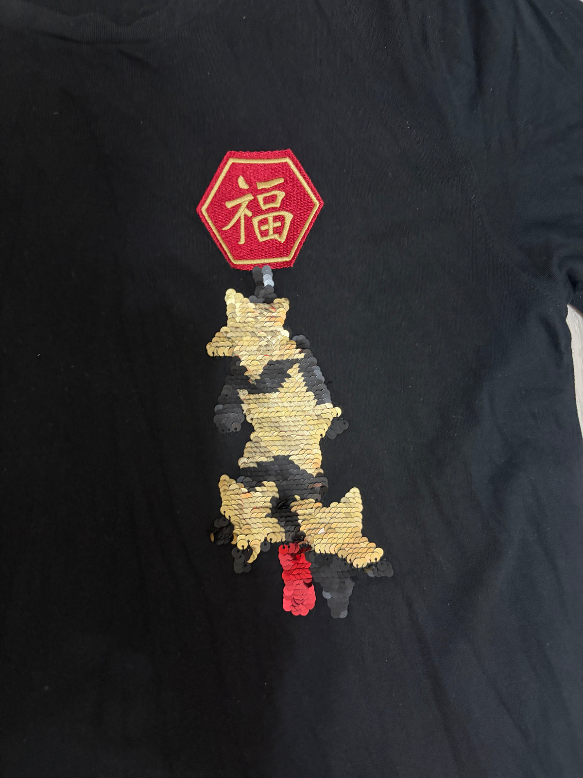 Close-up detail of the embroidered red hexagonal Chinese Fu character patch and gold sequin panda embroidery on a black t-shirt