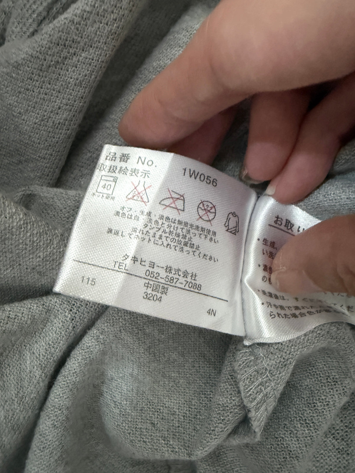 Close-up detail of the inner care label showing Takihyo distributor info, Made in China, product code 1W056, and wash instructions on a heather grey polo shirt
