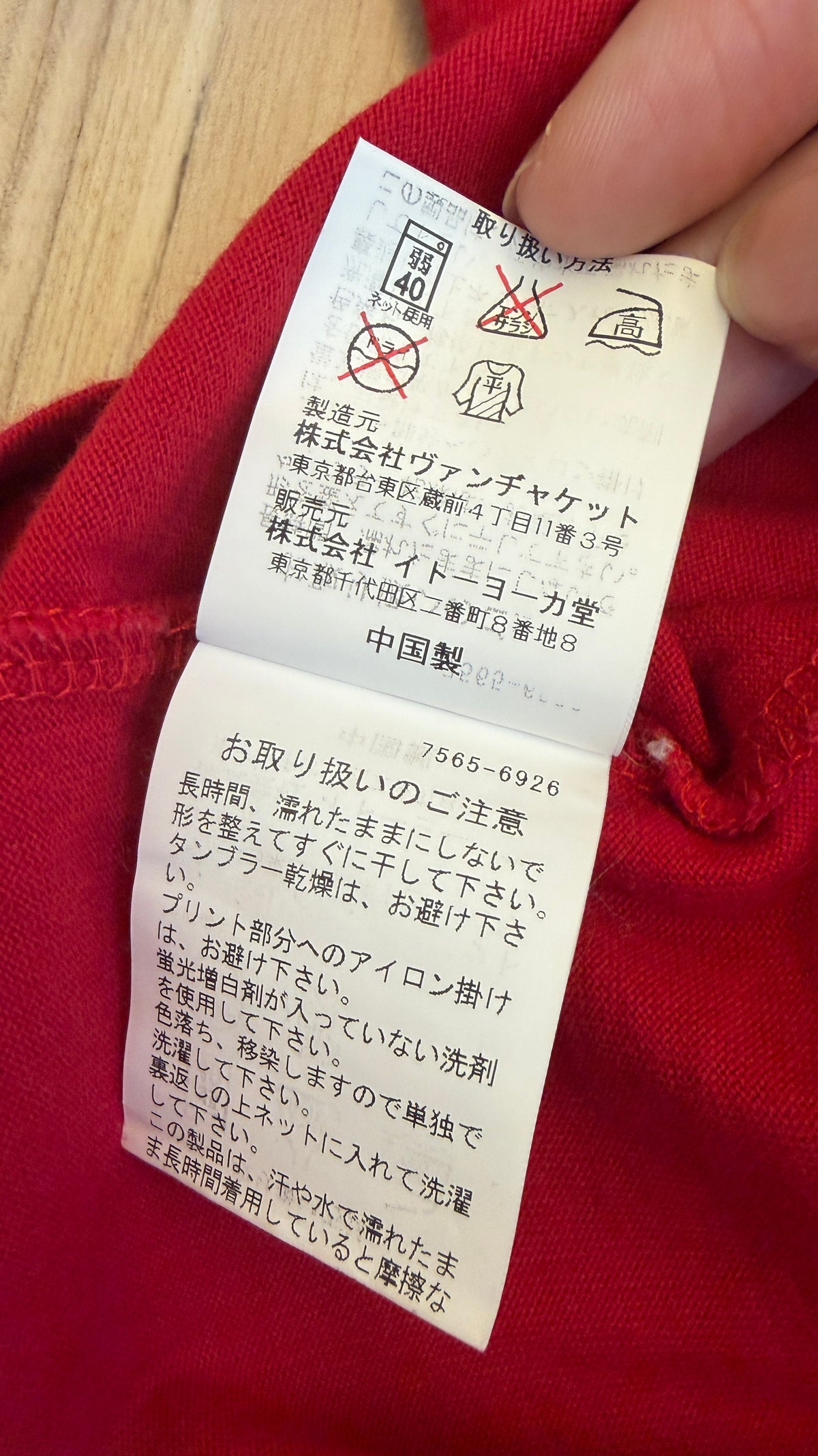 Close-up view of the inner white Japanese care instruction tag on a red t-shirt showing laundry symbols, manufacturer VAN Jacket Inc., distributor Ito-Yokado Co., Ltd., Made in China, and care notes
