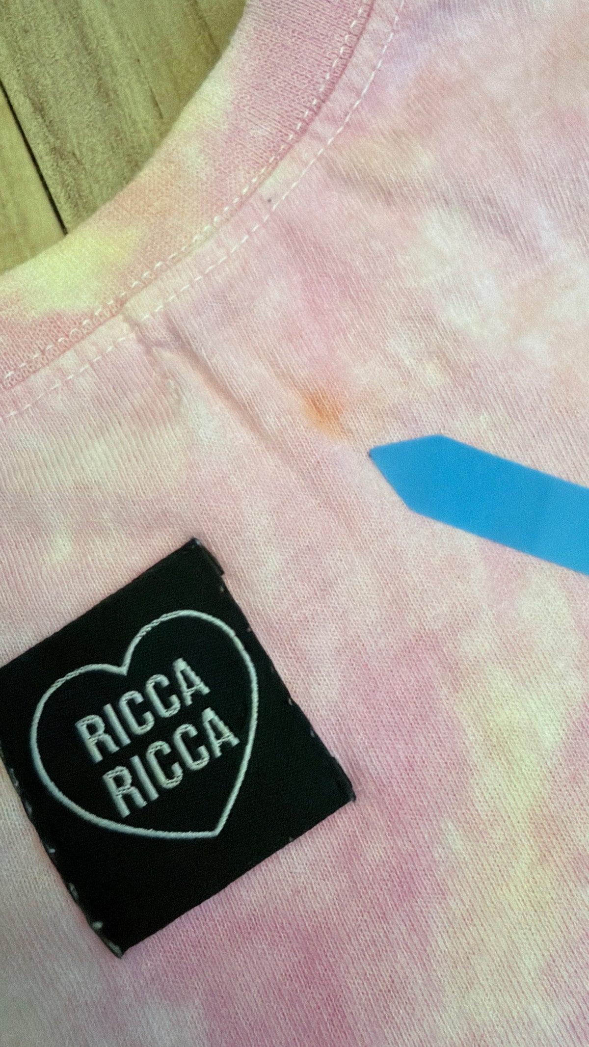 Close-up view of a pink tie-dye Ricca Ricca t-shirt showing a small orange stain defect pointed out by a blue sticker.