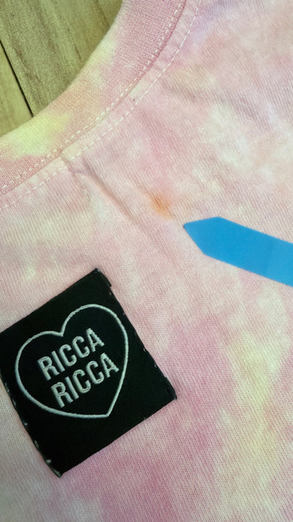 Close-up view of a pink tie-dye Ricca Ricca t-shirt showing a small orange stain defect pointed out by a blue sticker.