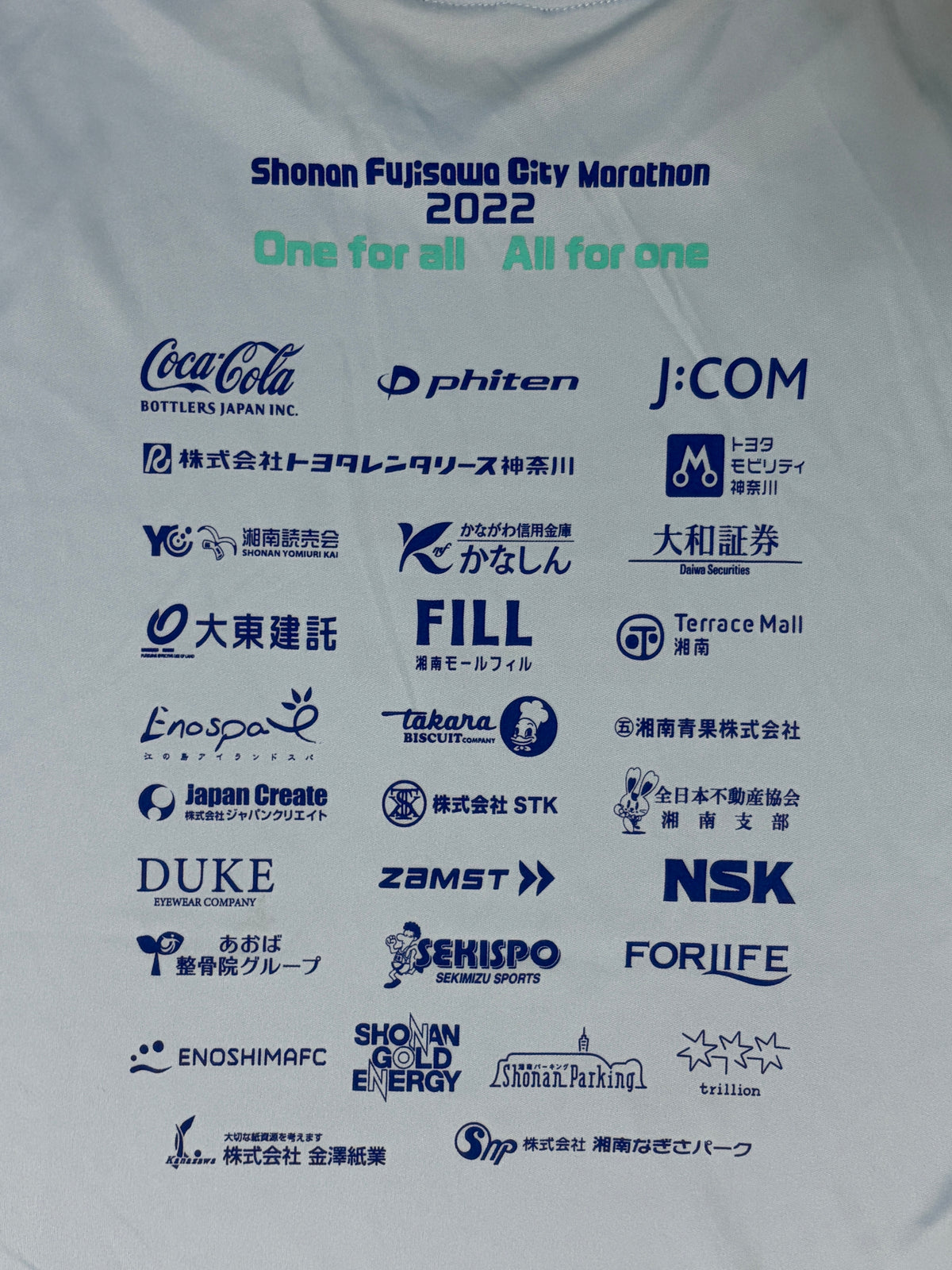 A close-up of a white athletic t-shirt printed with the Shonan Fujisawa City Marathon 2022 logo and numerous sponsor logos in blue and green text.