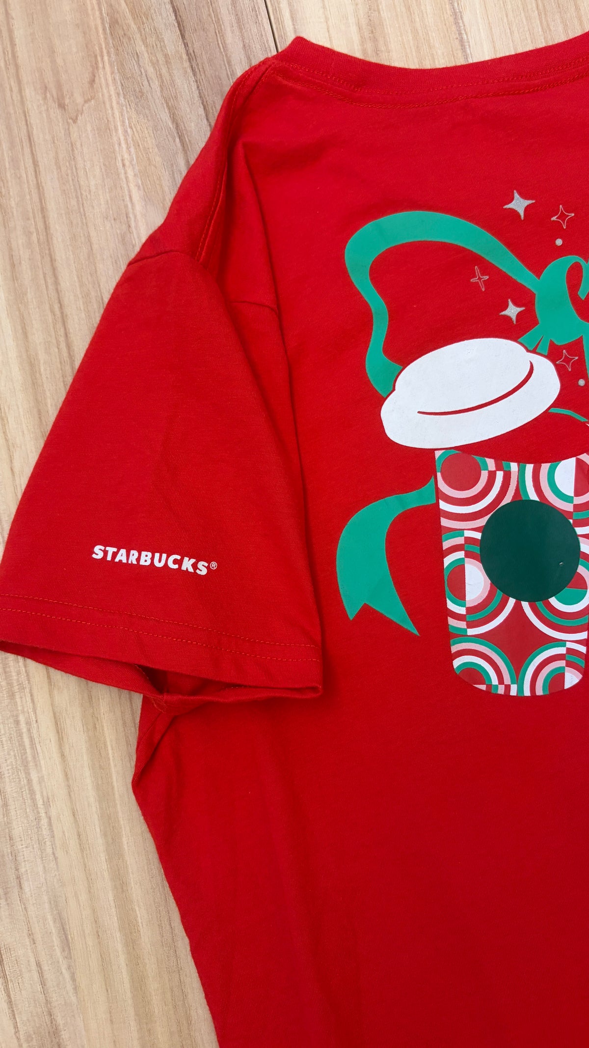 Close-up detail of a red Starbucks holiday t-shirt showing the white Starbucks logo printed on the sleeve alongside a festive holiday coffee cup graphic