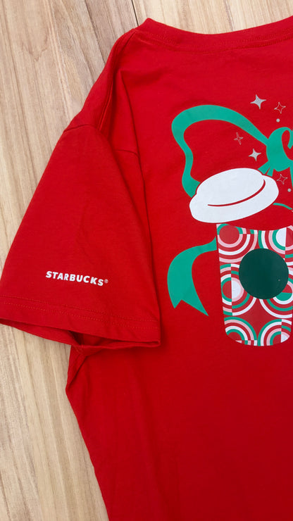 Close-up detail of a red Starbucks holiday t-shirt showing the white Starbucks logo printed on the sleeve alongside a festive holiday coffee cup graphic