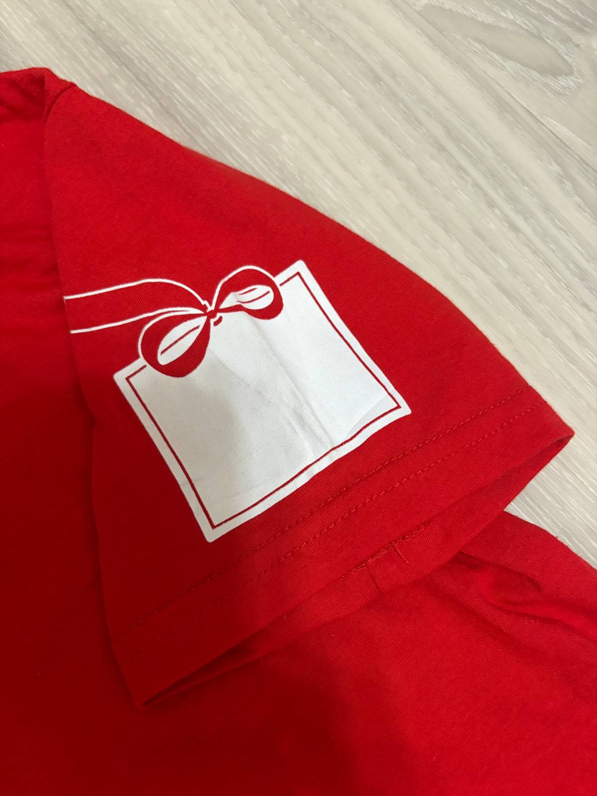 Close-up detail of the white ribbon gift tag graphic print on the short sleeve of a red Starbucks holiday t-shirt