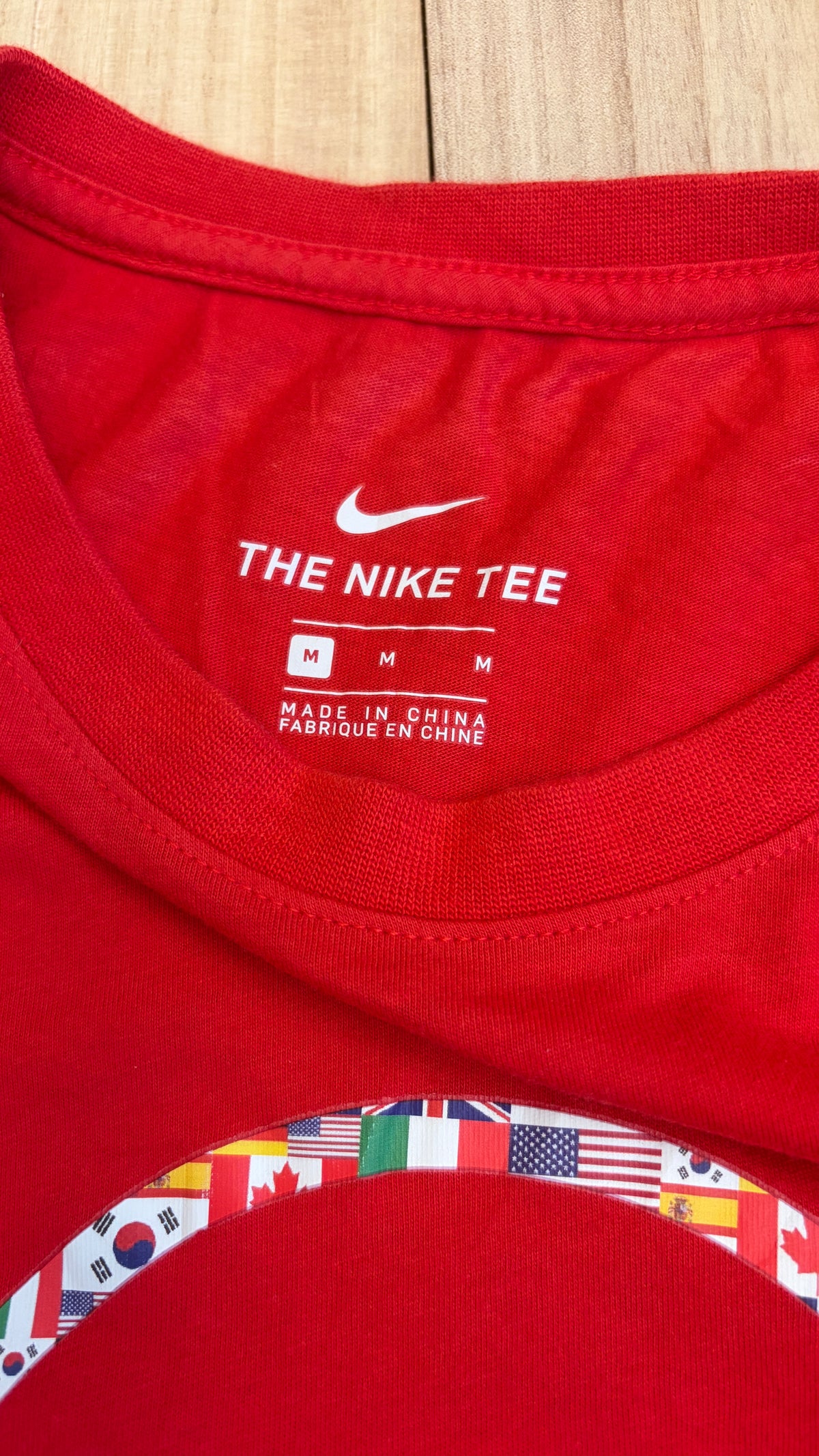 Close-up view of the inner collar of a red Nike t-shirt displaying a white Swoosh logo, The Nike Tee text, size M, and Made in China print above a world flag circle graphic