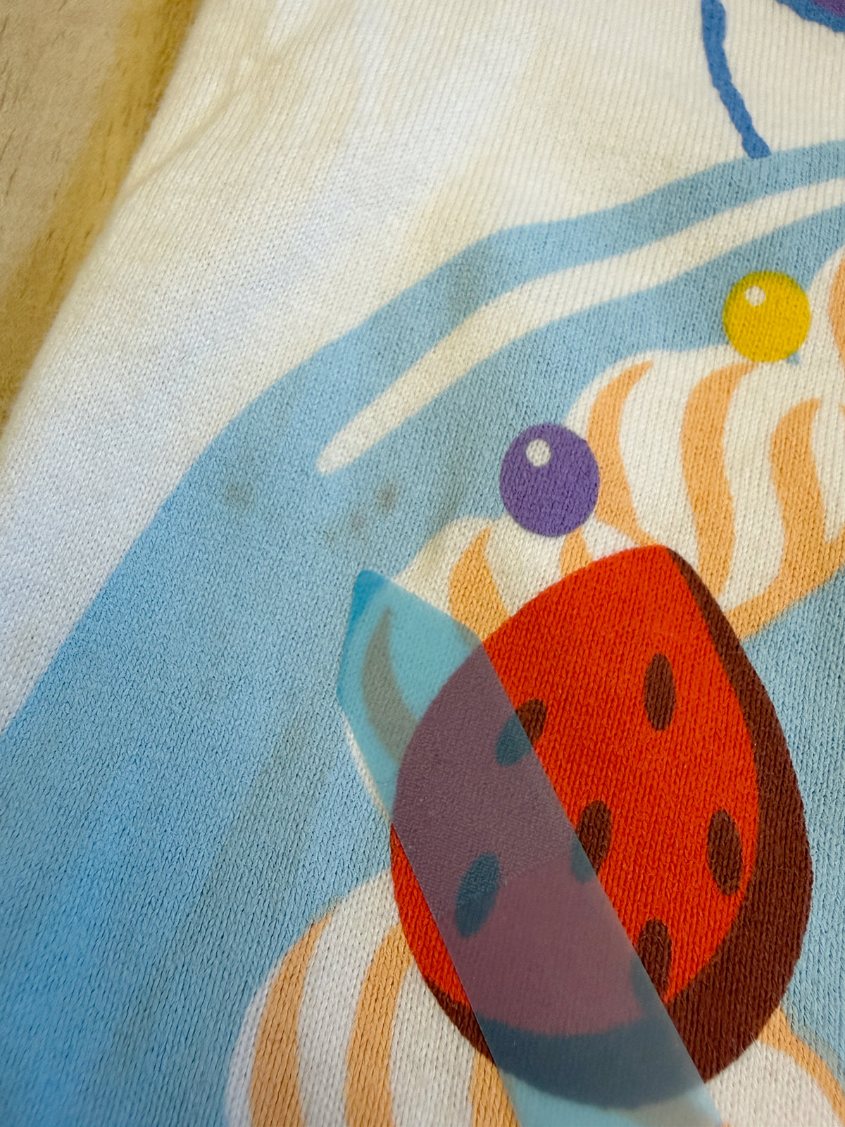 Close-up view of the cake print on a white Tokyo Disney Resort Its My Birthday graphic tee showing small stain defects pointed out by a sticker.