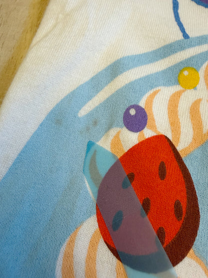 Close-up view of the cake print on a white Tokyo Disney Resort Its My Birthday graphic tee showing small stain defects pointed out by a sticker.